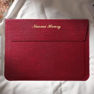 Puede incluir: Una funda roja para port&aacute;til con textura de piel de cocodrilo. El nombre "Naomi Henry" est&aacute; grabado en letras doradas en la parte delantera. La funda rectangular tiene un cierre de solapa y est&aacute; sobre una superficie blanca.
