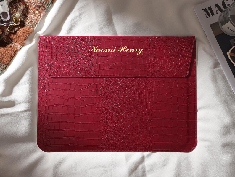 Puede incluir: Una funda roja para port&aacute;til con textura de piel de cocodrilo. El nombre "Naomi Henry" est&aacute; grabado en letras doradas en la parte delantera. La funda rectangular tiene un cierre de solapa y est&aacute; sobre una superficie blanca.