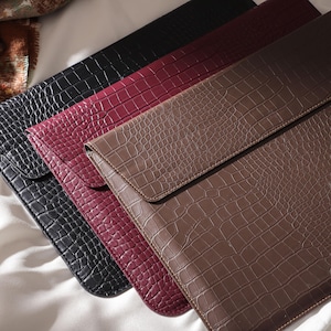 May include: Four leather laptop sleeves in a row, each with a different color and crocodile skin texture. The colors are black, burgundy, and brown. The sleeves are rectangular with a flap closure.