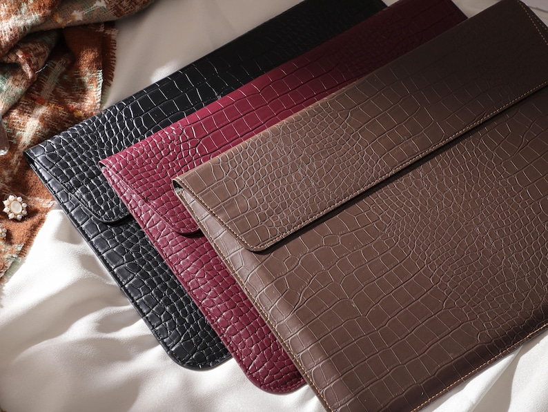 May include: Four leather laptop sleeves in a row, each with a different color and crocodile skin texture. The colors are black, burgundy, and brown. The sleeves are rectangular with a flap closure.