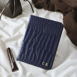 May include: A navy blue wallet with a crocodile skin texture and gold initials "C.K." is displayed on a white surface. A small tube of brown lip balm and a dark brown purse are also visible.