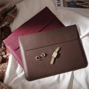 May include: A brown faux-leather laptop sleeve with a flap closure, textured to resemble crocodile skin, rests atop a burgundy sleeve. A gold watch, earrings, and a bracelet are arranged nearby. A scarf and perfume bottle are also visible.