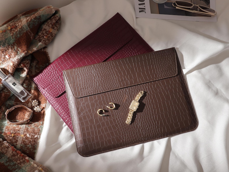 May include: A brown faux-leather laptop sleeve with a flap closure, textured to resemble crocodile skin, rests atop a burgundy sleeve. A gold watch, earrings, and a bracelet are arranged nearby. A scarf and perfume bottle are also visible.