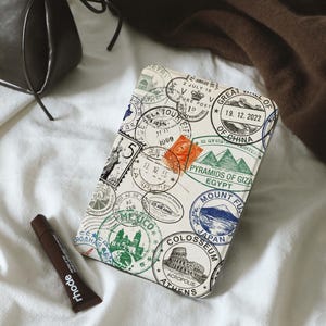 Puede incluir: Una bolsa crema con temática de viajes con varios sellos de todo el mundo, incluyendo la Torre Eiffel, el Coliseo y el Monte Fuji. Un tubo de producto labial está al lado de la bolsa. Un bolso y un suéter marrones están en el fondo.
