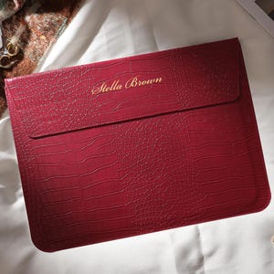 Puede incluir: Una funda para port&aacute;til burdeos con textura de piel de cocodrilo. Las palabras "Stella Brown" est&aacute;n impresas en dorado. La funda es rectangular con una solapa. Un reloj y pendientes dorados est&aacute;n al lado.