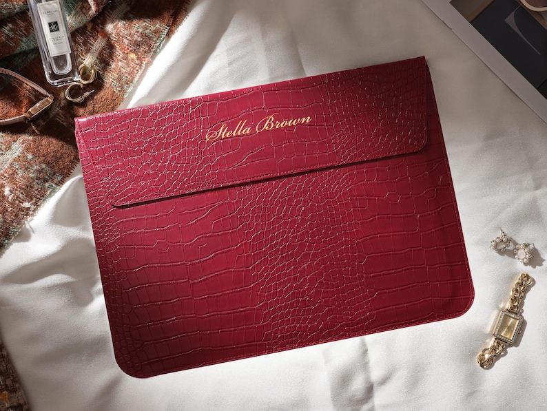 Puede incluir: Una funda para port&aacute;til burdeos con textura de piel de cocodrilo. Las palabras "Stella Brown" est&aacute;n impresas en dorado. La funda es rectangular con una solapa. Un reloj y pendientes dorados est&aacute;n al lado.