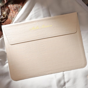 May include: Cream-colored laptop sleeve with a crocodile skin texture. The flap closure has the text "Hello Gorgeous" in gold lettering. The sleeve is on a white surface, with a gold watch and earrings nearby.