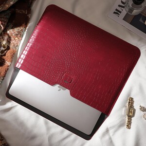 Puede incluir: Una funda roja para port&aacute;til con un patr&oacute;n texturizado similar a la piel de cocodrilo. La funda est&aacute; abierta, revelando un port&aacute;til plateado. Accesorios como un perfume, gafas de sol y joyas est&aacute;n dispuestos alrededor.