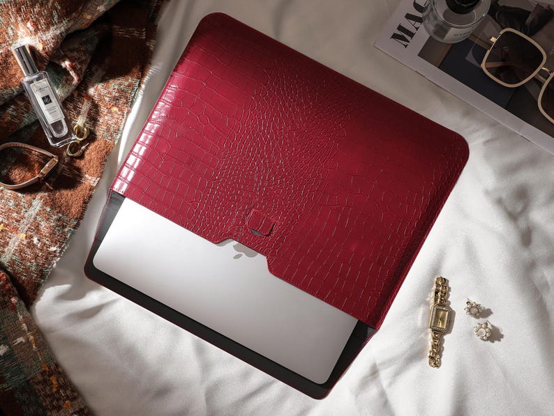 Puede incluir: Una funda roja para port&aacute;til con un patr&oacute;n texturizado similar a la piel de cocodrilo. La funda est&aacute; abierta, revelando un port&aacute;til plateado. Accesorios como un perfume, gafas de sol y joyas est&aacute;n dispuestos alrededor.