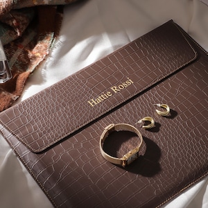 May include: A brown leather clutch with a crocodile texture, gold lettering that reads "Hattie Rossi", and a gold watch and earrings. A perfume bottle is visible in the background. The items are arranged on a white surface.