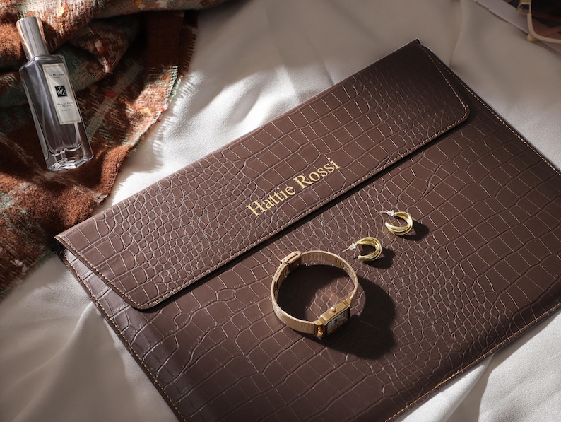 May include: A brown leather clutch with a crocodile texture, gold lettering that reads "Hattie Rossi", and a gold watch and earrings. A perfume bottle is visible in the background. The items are arranged on a white surface.