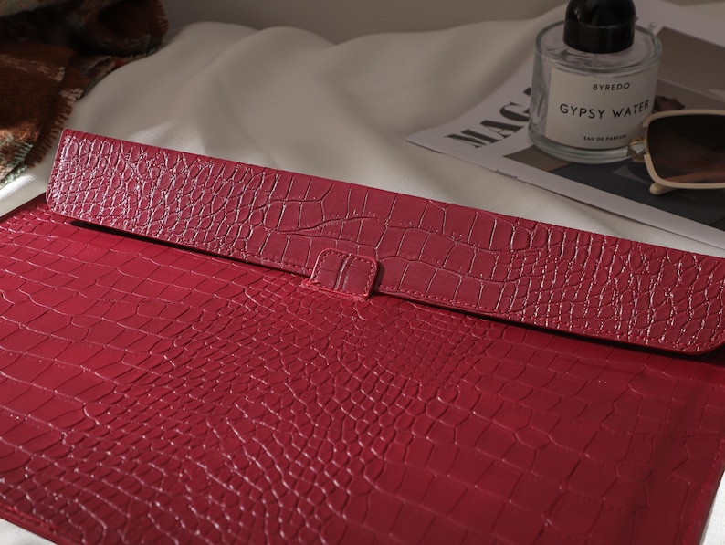 Puede incluir: Una funda roja para port&aacute;til con efecto piel de cocodrilo y cierre de solapa. La funda es de un tono rojo intenso y tiene una superficie texturizada. La solapa se asegura con una peque&ntilde;a leng&uuml;eta. La funda est&aacute; dise&ntilde;ada para proteger un port&aacute;til o una tableta.