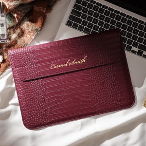 May include: A burgundy laptop sleeve with a crocodile texture, personalized with the name "Carmel Smith" in gold lettering. The sleeve is next to a laptop, a gold watch, and a perfume bottle.