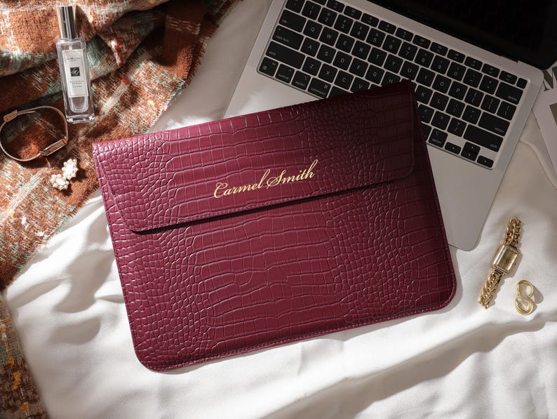 May include: A burgundy laptop sleeve with a crocodile texture, personalized with the name "Carmel Smith" in gold lettering. The sleeve is next to a laptop, a gold watch, and a perfume bottle.