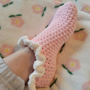 May include: A pink, crocheted slipper with a white, ruffled trim. The slipper is made of a soft, textured yarn and is designed to cover the foot. The slipper is resting on a white blanket with a pink floral pattern.