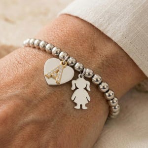 Può includere: Braccialetto con perline argentate con un ciondolo a forma di cuore con la lettera "A" e un piccolo ciondolo a forma di sagoma di ragazza. La lettera "A" è dorata e incastonata con piccole pietre trasparenti.