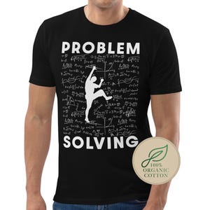 May include: Black t-shirt with the words "PROBLEM SOLVING" in white. A white silhouette of a person climbing is in the center. The background is filled with mathematical equations. A circular logo on the bottom right says "100% ORGANIC COTTON."