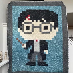 May include: A blue and gray quilt with a pixelated image of Harry Potter holding a wand. The quilt features a black background with a blue and white pattern.