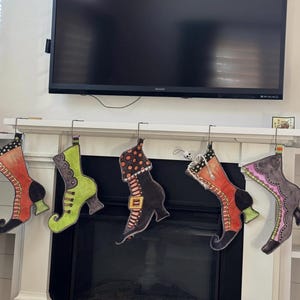 May include: Five Halloween-themed witch boot stockings hang from a white mantel. Each stocking is a different color and design, including orange, green, black, and purple. The boots have various embellishments and patterns, perfect for holiday decor.