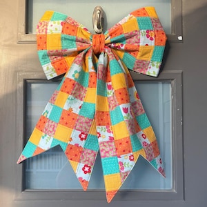 May include: A large decorative bow made of quilted fabric featuring a patchwork pattern of bright orange, teal, and yellow squares. The fabric includes various floral prints and small geometric designs, with two long, pointed tails hanging downward.