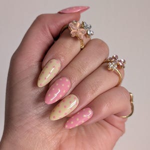 Puede incluir: Una mano que muestra u&ntilde;as en forma de almendra pintadas con una base amarilla clara y lunares rosas. La mano tambi&eacute;n lleva anillos dorados con dise&ntilde;os florales, con piedras preciosas rosas y transparentes. La u&ntilde;a del dedo me&ntilde;ique tambi&eacute;n est&aacute; pintada con lunares rosas.