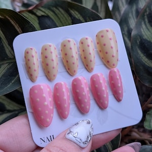 Puede incluir: Un juego de u&ntilde;as postizas en dos dise&ntilde;os. Uno es amarillo claro con lunares rosas, y el otro es rosa con lunares blancos. La palabra "NAIL" est&aacute; impresa en la tarjeta.