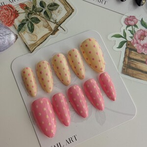 Puede incluir: Un conjunto de u&ntilde;as postizas de varios tama&ntilde;os, con base amarilla clara con lunares rosas y base rosa con lunares amarillo claro. Las palabras "NAIL ART" est&aacute;n impresas en la tarjeta.