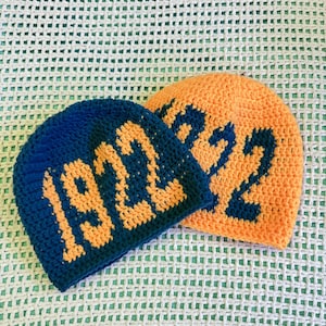 Könnte beinhalten: Zwei gehäkelte Mützen. Eine ist marineblau mit orangefarbenen Buchstaben "1922", und die andere ist orange mit marineblauen Buchstaben "1922". Die Mützen werden auf einer weiß-grünen gehäkelten Oberfläche präsentiert.