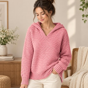 May include: A pink long-sleeved hooded sweater featuring a textured crochet knit pattern, a V-neckline, and ribbed cuffs and hem. The garment is constructed with a thick yarn and has a relaxed fit, providing a consistent solid color throughout.