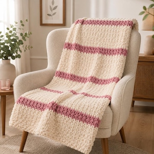 May include: A chunky knit throw blanket featuring a repeating pattern of wide cream-colored sections and narrower horizontal stripes in a dusty rose pink color. The blanket has a thick, textured yarn construction with a soft, plush appearance.