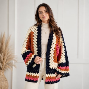 May include: A crochet cardigan featuring a granny square pattern in navy blue, cream, rust orange, and bright pink. The open-front garment has long, wide sleeves and a straight hem, worn over a cream-colored turtleneck sweater with a simple gold necklace.