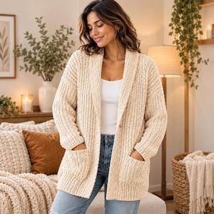 Peut inclure: Un cardigan en tricot épais de couleur crème avec un col châle, des manches longues et deux poches plaquées à l'avant. Ce gilet ouvert présente une texture côtelée verticale et est porté sur un débardeur blanc et un jean bleu.