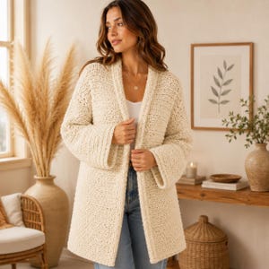 May include: A cream-coloured, long-sleeved, open-front cardigan featuring a textured, chunky knit pattern. The garment has a wide shawl collar, straight hemline, and loose-fitting sleeves, worn over a simple white top and blue denim jeans.