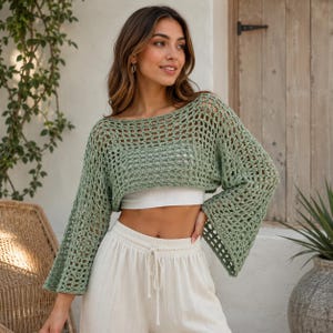 May include: A sage green crochet sweater featuring a wide, open-knit mesh pattern, a boat neckline, and long, flared bell sleeves. The garment is cropped in length and worn over a plain white bandeau top, paired with white drawstring trousers.