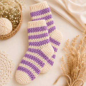 Könnte beinhalten: Ein Paar handgehäkelte Socken mit horizontalen Streifen in abwechselnden Farbtönen von Helllila und Creme. Die Socken verfügen über ein geripptes Bündchen, einen verstärkten Fersenbereich und eine abgerundete Spitze aus strukturiertem Garn.