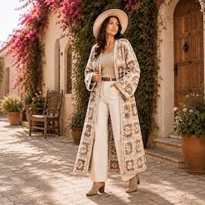 Könnte beinhalten: Ein langer, vorne offener Duster-Cardigan mit einem beigefarbenen und braunen Häkelmuster aus Granny Squares, getragen über einem hellbeigen Tanktop und einer weißen Hose mit braunem Gürtel, kombiniert mit einem beigen Hut und Stiefeletten.
