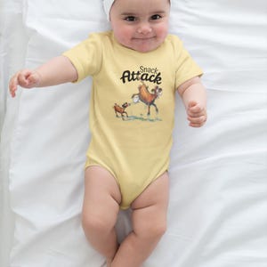 Op de afbeelding: Een lichtgele babybody met de tekst "Snack Attack" en een cartoonillustratie van een hert en een kleiner dier. De baby draagt een witte haarband en ligt op een wit laken.