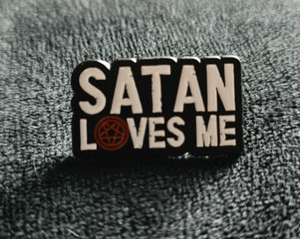 May include: A black enamel pin featuring the white text SATAN LOVES ME, with the letter O in LOVES replaced by a red circular pentagram symbol. The pin has a rectangular shape with rounded edges and a bold, high-contrast design.