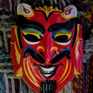 Puede incluir: Una máscara roja vibrante con una expresión diabólica. La máscara presenta detalles en negro, amarillo, azul y blanco, incluyendo cuernos, cejas y perilla. El fondo es una mezcla texturizada de telas y cuerdas coloridas.