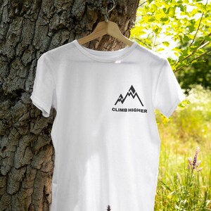 May include: White t-shirt with a black mountain graphic and the words "CLIMB HIGHER" printed below. The shirt is hanging on a wooden hanger against a tree trunk and green foliage.