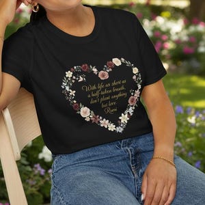 Könnte beinhalten: Schwarzes T-Shirt mit floralem Herzdesign und dem Zitat "Mit dem Leben so kurz wie ein halbgeatmeter Atemzug, pflanze nichts als Liebe." Das Design umfasst bordeauxrote und weiße Blumen. Das Model trägt eine blaue Jeans.