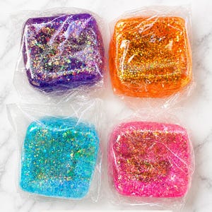 May include: Four 1.9-inch cube squishies wrapped in clear plastic. The set includes one purple, one orange, one blue, and one pink cube, each filled with multi-colored glitter particles that reflect light. The text 1.9" CUBE SQUISHY is visible.