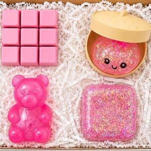 May include: A set of items including a pink nine-square wax melt block, a round beige container with a lid holding a pink glittery slime with a smiling face, a pink translucent bear-shaped figure, and a square container filled with pink glitter.