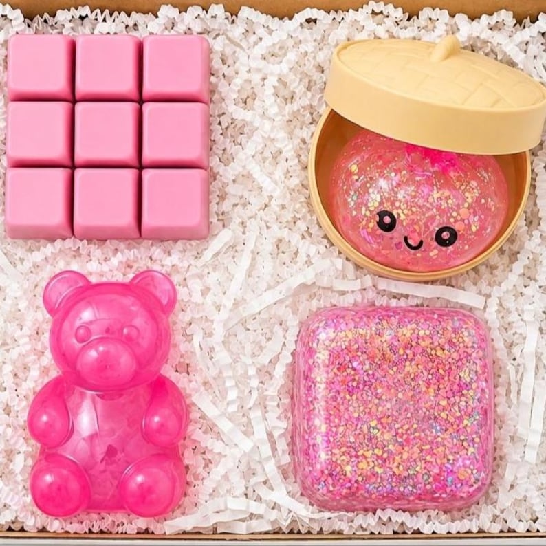 May include: A set of items including a pink nine-square wax melt block, a round beige container with a lid holding a pink glittery slime with a smiling face, a pink translucent bear-shaped figure, and a square container filled with pink glitter.