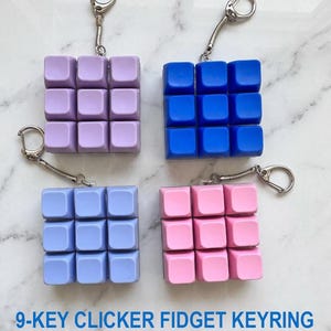 May include: Four square 9-key clicker fidget keyrings in lavender, blue, periwinkle, and pink. Each keyring features a grid of nine square buttons attached to a silver metal split ring by a short chain. The text 9-KEY CLICKER FIDGET KEYRING is visible.