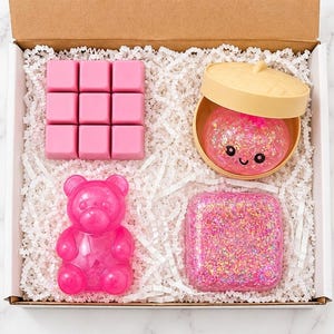 May include: A gift box containing four items: a pink nine-cube grid, a beige lidded container holding a pink glittery sphere with a smiling face, a translucent pink bear-shaped figure, and a square block filled with pink and gold glitter.