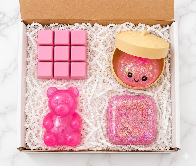May include: A gift box containing four items: a pink nine-cube grid, a beige lidded container holding a pink glittery sphere with a smiling face, a translucent pink bear-shaped figure, and a square block filled with pink and gold glitter.