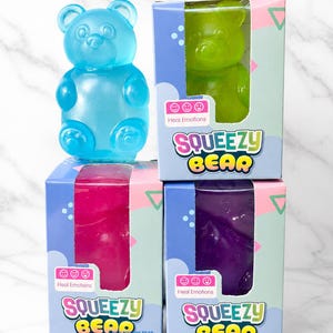 May include: A blue translucent bear-shaped toy stands next to three rectangular boxes labeled SQUEEZY BEAR and Heal Emotions. The boxes contain green, pink, and purple bear-shaped toys. The text 3 INCH SQUISHY BEAR is printed at the bottom.