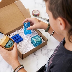 May include: A cardboard gift box filled with white paper shred containing a blue translucent gummy bear figure, a blue square fidget toy, a blue glittery cube, and a small round container holding a glittery sphere with a printed face.