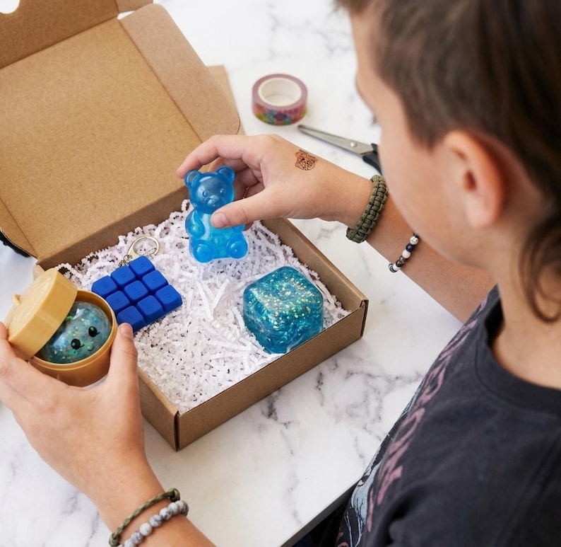 May include: A cardboard gift box filled with white paper shred containing a blue translucent gummy bear figure, a blue square fidget toy, a blue glittery cube, and a small round container holding a glittery sphere with a printed face.